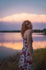 Giovane donna in abito estivo in piedi sulla riva del lago al tramonto — Foto stock