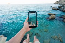 Hand eines Mannes, der ein Foto mit dem Smartphone von Beinen macht, während er auf einer Klippe über dem türkisfarbenen Meer sitzt — Stockfoto