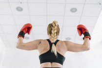 Vista posterior de la mujer musculosa con guantes de boxeo rojos mostrando bíceps mientras está de pie sobre el fondo borroso del gimnasio - foto de stock
