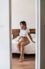 Приваблива татуйована жінка в трусиках і футболці сидить на ліжку і дивиться вниз — стокове фото