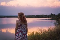 Romantic woman in summer dress standing on lake shore at sunset — Stock Photo