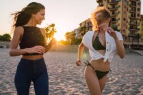 Lachende Freundinnen laufen am Strand mit Gebäuden im Hintergrund — Stockfoto