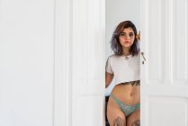 Молодая татуированная женщина в трусиках и футболке стоит в дверях и смотрит в камеру дома — стоковое фото