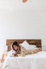Молодая женщина в шелковом халате лежит на кровати и делает наброски в блокноте в стильной спальне — стоковое фото