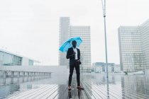 Homme d'affaires ethnique tenant un parapluie bleu debout sur un trottoir mouillé en ville — Photo de stock