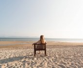 Rückansicht einer Frau, die es sich auf einem Stuhl am Sandstrand gemütlich macht und auf das Meer blickt — Stockfoto