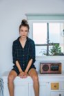 Attractive young woman in oversize checkered shirt sitting on counter near retro radio receiver and looking at camera — Stock Photo