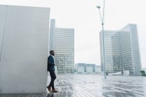 Homme d'affaires afro-américain appuyé sur le mur à l'extérieur avec des bâtiments modernes sur le fond — Photo de stock