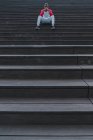 Homme ethnique en vêtements de sport assis sur des escaliers gris humides et la tête d'appui avec les mains pliées et regardant la caméra — Photo de stock