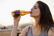 Счастливая молодая женщина пьет пиво из бутылки на открытом воздухе — стоковое фото