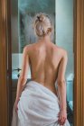 Вид ззаду молода жінка, загорнута в білий рушник, стоячи в дверях у ванній після душу — стокове фото