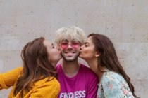 Mujeres jóvenes besándose feliz hombre rubio en camiseta rosa y gafas de sol sobre fondo texturizado gris - foto de stock