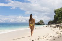 Vue arrière de la femme en bikini jaune marchant sur la plage de sable fin à l'océan — Photo de stock