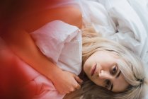 Молода жінка приймає м'яке покривало і дивиться на камеру, лежачи на ліжку — стокове фото