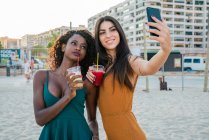 Mujeres multiétnicas de moda tomando bebidas y tomando selfie mientras con el teléfono inteligente en la playa - foto de stock