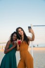 Модні багатоетнічні жінки п'ють і приймають селфі під час перебування зі смартфоном на пляжі — стокове фото