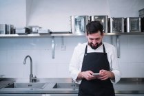 Side view of young man in cook uniform leaning on kitchen counter and using modern smartphone while standing in restaurant kitchen — Stock Photo