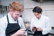 From below shot of two guys in cook uniform standing on restaurant kitchen and browsing smartphones during break — Stock Photo
