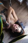 Чуттєва татуйована жінка, що покриває груди і торкається шиї, лежачи на дерев'яній підлозі — стокове фото
