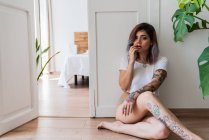 Donna scalza con tatuaggi toccare le labbra e guardando la fotocamera mentre seduto sul pavimento vicino alla porta della camera da letto elegante — Foto stock
