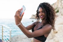 Весела красива чорна жінка бере селфі з мобільним телефоном на узбережжі — стокове фото