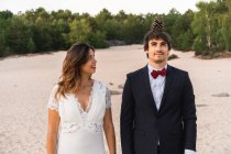 Lustiger Bräutigam mit Tannenzapfen auf dem Kopf und ausdrucksstark überraschter Braut, die zusammen an der Küste steht und in die Kamera schaut — Stockfoto