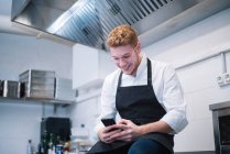 Вид сбоку на молодого человека в форме повара, опирающегося на кухонный прилавок и использующего современный смартфон, стоя на кухне ресторана — стоковое фото