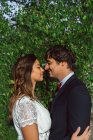 Side view of bride and groom in love looking at each other happily while standing in lush green foliage in sunlight — Stock Photo