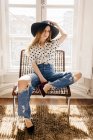 Stylische junge Frau mit Hut sitzt auf Stuhl und blickt in die Kamera — Stockfoto