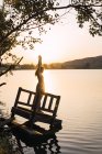 Frau im Kleid mit erhobenen Händen auf versunkenem Holzsteg am See bei Sonnenuntergang — Stockfoto