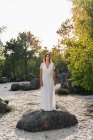 Adult woman wearing elegant lace white dress of bride and standing on rock on sandy coastline in green foliage looking at camera — Stock Photo