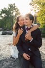 Glücklich elegante Männer und Frauen in Hochzeitsoutfits, die sich am Strand umarmen und im Sonnenlicht lächelnd wegschauen — Stockfoto