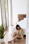 Giovane donna in abito di seta seduta sul pavimento e disegno in sketchbook in camera da letto elegante — Foto stock