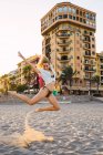 Flexible young woman jumping on beach with buildings on background — Stock Photo