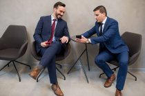 Dos hombres elegantes vistiendo trajes y sentados en sillas en la oficina compartiendo teléfono inteligente y divirtiéndose en el ocio - foto de stock