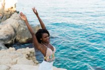 Elegant black woman standing on cliff with hands up above water — Stock Photo