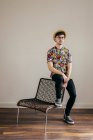 Stilvoller junger Mann mit Strohhut und gemustertem Hemd sitzt auf Stuhl gegen graue Wand — Stockfoto