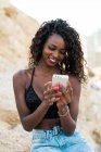 Charming young curly woman using smartphone while sitting on rock — Stock Photo