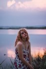 Jeune femme tendre en robe florale debout sur la rive du lac au coucher du soleil et regardant la caméra — Photo de stock