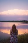 Woman in summer dress standing on lake shore at sunset — Stock Photo
