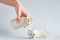 Человеческая рука, держащая белый керамический кувшин и наливая воду в чашку с цветочным орнаментом на белом фоне с белой орхидеей — стоковое фото