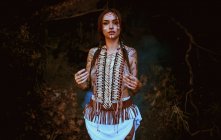 Attraente giovane donna con dipinti tradizionali indiani sul viso guardando la fotocamera e in piedi nella foresta — Foto stock