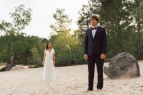 Hombre y mujer en elegantes vestidos de novia de pie por separado en la costa de arena con árboles verdes a la luz del sol en el fondo - foto de stock