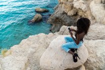 Stylish black woman in heels and jeans sitting on rock above sea water and using mobile phone — Stock Photo