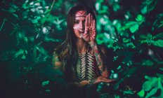 Jolie jeune femme avec des peintures indiennes traditionnelles sur le visage regardant la caméra et debout dans la forêt — Photo de stock