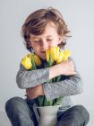 Jeune garçon avec les yeux fermés assis et embrassant bouquet de tulipes jaunes sur fond gris — Photo de stock