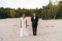 Funny groom with pine cone on head and expressive surprised bride standing together on coast looking at camera — Stock Photo