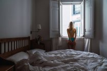 Donna bionda senza maglietta che guarda fuori dalla finestra in camera da letto — Foto stock