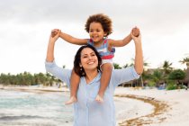Cheerful ethnic woman holding and carrying little son on shoulders while walking on sandy coast at seaside — Stock Photo