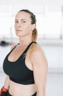 Confident woman in sportswear standing on blurred background of gym and looking at camera — Stock Photo